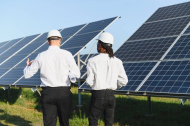Güneş enerjisi tarlasında dikilen kasklı ve beyaz gömlekli mühendisler yenilenebilir enerji üretimini tartışıyorlar.