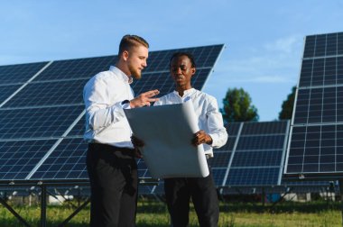 Tasarım planlarını inceleyen iki mühendis güneş panellerinin arasında duruyor. Yenilenebilir enerji gelişimi üzerine yoğunlaşmak