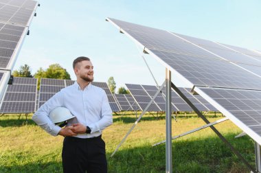 Genç erkek mühendis kaskını tutuyor, açık mavi gökyüzü ile büyük bir güneş paneli alanında duruyor.