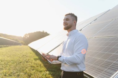 Genç adam, güneş panelinin verimliliğini dijital tabletle izliyor, yenilenebilir yeşil enerji projesi üzerinde çalışıyor