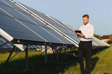 Güneş enerjisi santralinin performansını izleyen mühendis, güneş panellerinin yanında dijital bir tablet tutuyor.