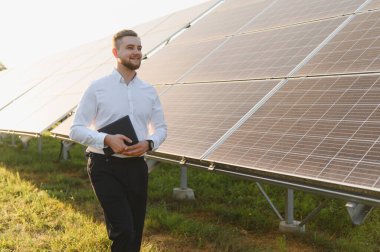 Elinde tablet olan gülümseyen mühendis, bir enerji santralinde güneş panellerinin önünde duruyor, sürdürülebilir enerji planlıyor.