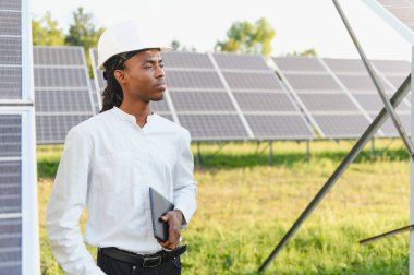 Siyah şapkalı Afrikalı Amerikalı mühendis elinde dijital tablet, güneş paneli dizilerinin yanında duruyor.