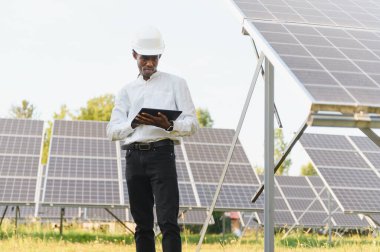 Güneş paneli sahasında duran, dijital tablet tutan ve denetleme sistemini kontrol eden genç siyah adam mühendisi.