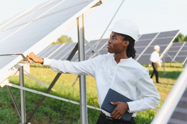 Afrika kökenli Amerikalı mühendis sabit şapka takıyor ve elinde tablet, güneş panellerini kontrol ediyor, sürdürülebilir enerji üretimini sağlıyor.
