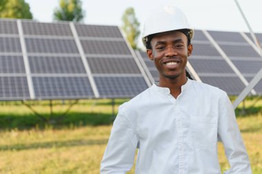 Afrikalı erkek mühendis, sert bir şapka takıyor, gülümsüyor, güneş panellerinin önünde duruyor, yenilenebilir enerjiyi temsil ediyor.