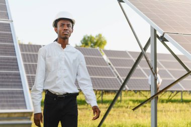 Genç zenci teknisyen, yenilenebilir enerji santralinde güneş panellerini inceliyor.