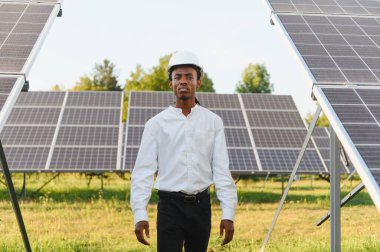 Beyaz bir şapka ve gömlek giyen genç siyah mühendis yeşil bir alanda güneş panellerinin arasında duruyor.