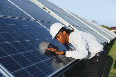 Büyük güneş panellerini denetleyen, sürdürülebilir temiz enerji üretimi ve verimli faaliyet sağlayan sert şapkalı Afrikalı Amerikalı bir adam.