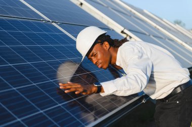 Güneş panellerini denetleyen kasklı genç bir adam. Temiz enerji teknolojisi ve sürdürülebilir güç üretimi vurgulanıyor