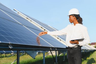 Kask takan ve elinde tablet tutan genç bir adam yenilenebilir enerji alanındaki güneş panelinin yüzeyini kontrol ediyor.