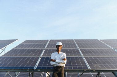 Afrikalı Amerikalı bir mühendis, güneş panelleri önünde kollarını kavuşturup gülümsüyor, temiz enerjiyi teşvik ediyor.