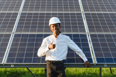 Şapka takan ve gülümseyen genç bir adam, baş parmak işareti yapıyor, güneş panelinin önünde duruyor.