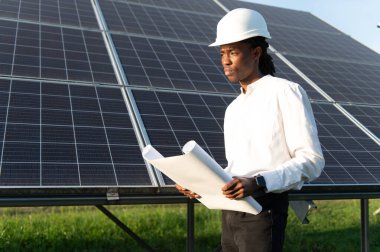 Sert şapka takan genç bir adam planlar tutuyor, güneş panellerinin yanında duruyor, açık havada yenilenebilir enerji projesi üzerinde çalışıyor.