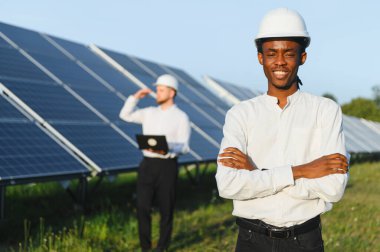 Bir fotovoltaik enerji santralinde meslektaşımla güneş enerjisi projesinde çalışırken kameraya gülümseyen genç bir adam.