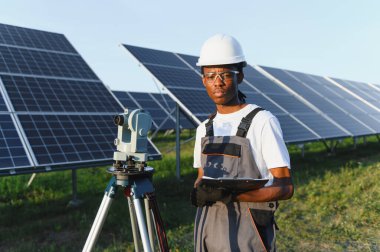 Afrika kökenli Amerikalı mühendis araştırma ekipmanlarıyla duruyor, elinde bir dosya var, bir güneş enerjisi tesisinde çalışıyor.