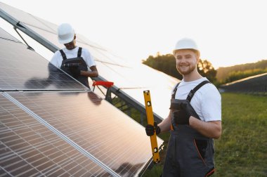 İki mühendis bir güneş çiftliğinde fotovoltaik paneller kuruyorlar. Biri gülümsüyor, baş parmak işareti yapıyor.