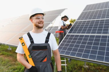 Gülümseyen erkek mühendis elinde bir aletle bir güneş enerjisi santralinin önünde duruyor. Bir meslektaşım arka plandaki paneller üzerinde çalışıyor.