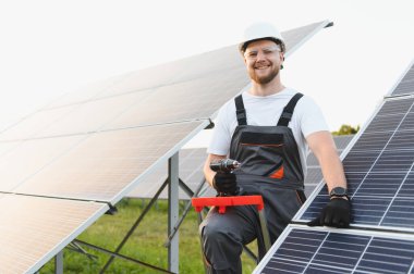Kask ve güvenlik gözlüğü takan gülümseyen teknisyen, güneş panelleri kuruyor, temiz enerji ve sürdürülebilirliğe katkıda bulunuyor