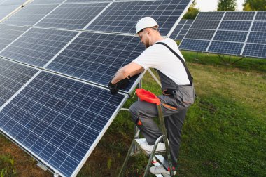 Sert şapkalı ve güvenlik gözlüklü erkek güneş teknisyeni güneşli yeşil bir alanda fotovoltaik paneller ve sürdürülebilir enerji çalışmaları sürdürüyor.