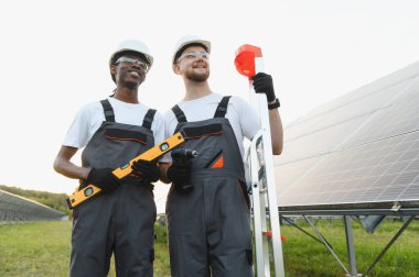 Güneş panellerini güneşli, sürdürülebilir bir güneş enerjisi santraline kuran kasklı ve güvenlik teçhizatlı iki gülümseyen etnik adam.