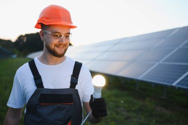 Genç mühendis gülümsüyor, elinde bir ampul tutuyor, güneş panellerinin önünde duruyor. Yenilenebilir enerji ve yenilik kavramı