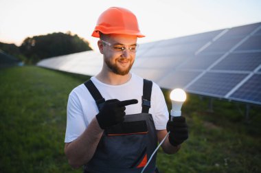 Güneş paneli çiftliğinde duran ve temiz enerji yeniliklerini temsil eden parlayan ampulü tutan güvenlik donanımı işçisi.