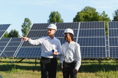 Çok ırklı mühendisler sert şapkalar takıyor, yenilenebilir enerji kurulumunu tartışıyor, güneş panellerinin önünde duruyorlar.