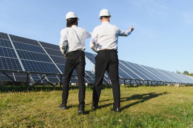 Kasklı ve planlı iki mühendis güneşli bir günde büyük bir güneş paneli tesisinde sürdürülebilir enerjiyi tartışıyor.