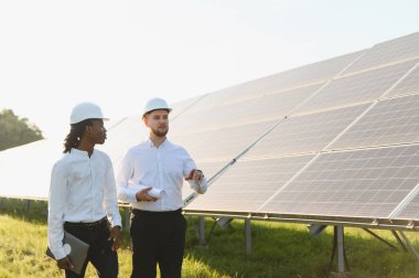 İki mühendis, biri siyah, biri beyaz. Büyük bir güneş paneli tesisini inceliyorlar. Yenilenebilir enerji projesi planlarını tartışıyorlar.
