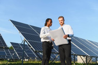 Çeşitli mühendisler bir güneş enerjisi santralinin planlarını inceliyor, takım çalışmasını, yeniliği ve sürdürülebilir enerji gelişimini sembolize ediyor