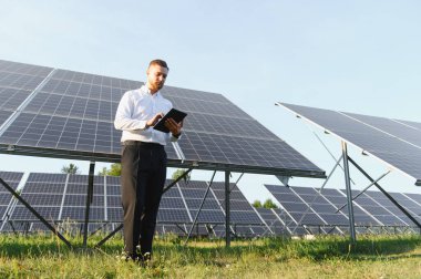 Gündelik kıyafet işindeki adam yenilenebilir enerji yönetimi için dijital tablet kullanarak güneş paneli alanını inceliyor.