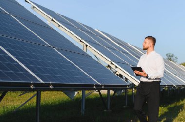 Bir dizi güneş panelini inceleyen genç erkek mühendis, performans ve verimliliği izlemek için dijital tablet tutuyor.