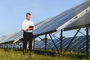 Beyaz gömlekli bir adam güneş enerjisi santralinde çalışıyor. Teftiş ve temiz enerji yönetimi için tablet kullanıyor.