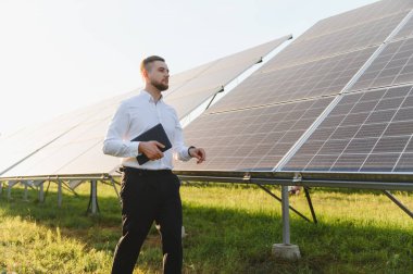 Güneş panelleri arasında yürüyen ve sürdürülebilir bir enerji santralinde enerji üretimini izleyen bir tablet tutan erkek mühendis.