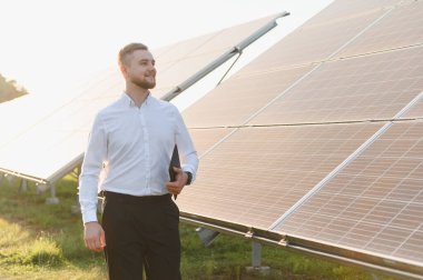 Genç işadamı güneş panellerinin önünde dururken elinde bir tablet tutarak yenilenebilir enerji çözümleri sunuyor.