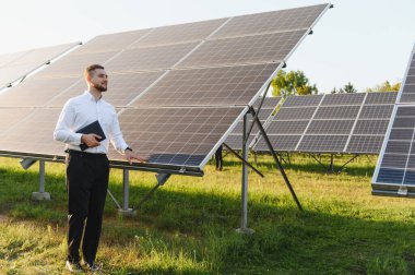Genç adam güneş panellerinin yanında duran tableti tutuyor, gelecekte yenilenebilir temiz enerji çözümleri ve sürdürülebilirlik geliştiriyor.