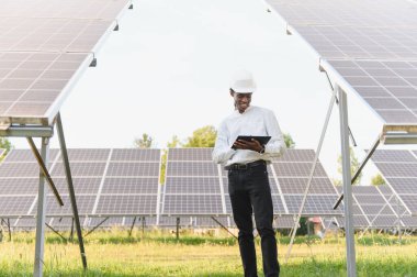 Büyük bir güneş çiftliğindeki fotovoltaik modülleri incelemek için bir tablet kullanan sert şapkalı Afrikalı Amerikalı mühendis. Yeşil enerji kavramı