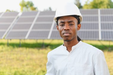 Siyah mühendis sabit bir şapka takıyor, güneş panellerinin önünde güvenle duruyor, yenilenebilir enerji ve sürdürülebilirliği temsil ediyor.