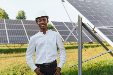 Genç mühendis gülümsüyor, güneş panelleri olan bir alanda sert bir şapka takıyor, yenilenebilir enerji ve sürdürülebilirliği temsil ediyor.