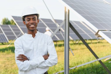 Sert bir şapka ve beyaz gömlek giyen Afrikalı mühendis güneş paneli alanında çapraz kollarıyla durarak yenilenebilir enerji yüklüyor.