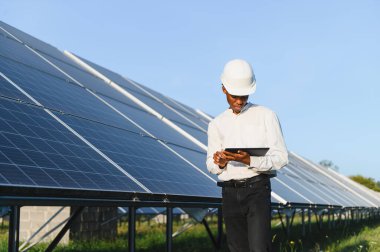 Kara şapkalı Afrikalı Amerikalı mühendis, veri takibi için dijital tablet kullanarak büyük güneş panellerini denetliyor.