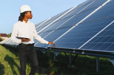 Siyah şapkalı ve tabletli Afro-Amerikan mühendis güneş panellerini inceliyor, sürdürülebilir enerji ve yenilenebilir teknolojiyi temsil ediyor.