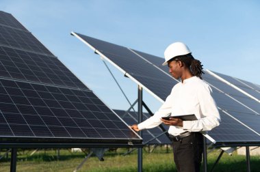 Sert şapkalı ve tabletli erkek mühendis güneş panellerini kontrol ederek bir enerji santralinde verimli bir yenilenebilir enerji operasyonu sağlıyor.