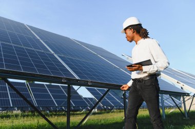 Sert bir şapka takan Afrikalı mühendis elinde dijital tablet, dışarıdaki fotovoltaik güneş panellerini inceliyor. Sürdürülebilir enerji kavramı