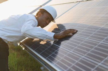 Sert bir şapka takan Afrikalı erkek mühendis, bir elektrik santralinde güneş panellerini denetliyor, yenilenebilir temiz enerjiyi teşvik ediyor.