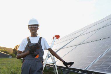 Güneş panelleri yakınında elinde güç aleti tutan, şapkalı ve gözlüklü gülümseyen adam, yenilenebilir enerji kurulumu üzerinde çalışıyor.
