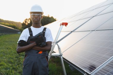 Bir elektrik santralinde güneş panelleri önünde çapraz kollarıyla matkap tutan genç siyah adam teknisyeni.