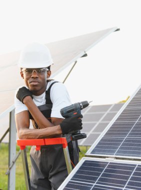 Afrikalı teknisyen güneş panelleri kuruyor, matkap tutuyor, başlık takıyor ve güvenlik gözlüğü takıyor, yenilenebilir enerji sağlıyor.