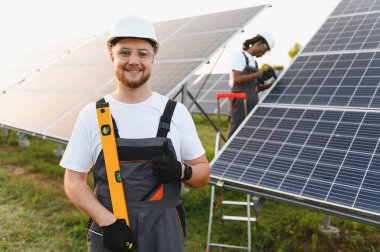 Bir güneş çiftliğinde, temiz enerji kurulumunu ve sürdürülebilir kalkınmayı sembolize eden bir aracı tutan gülen işçi